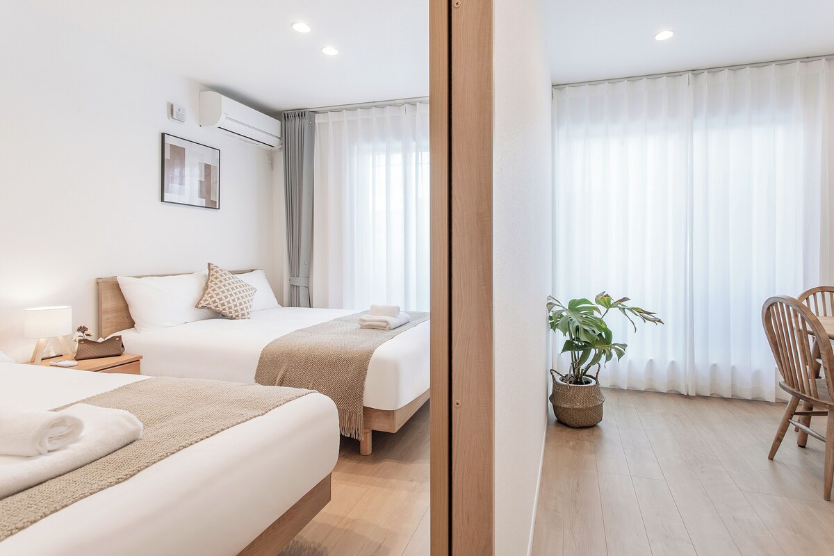 A hotel room with two beds, a bedside table with a lamp, a potted plant, and a view of a balcony with curtains.