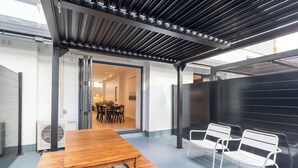A covered outdoor seating area with a table and chairs, an open doorway leading to an interior space, and a black wall.