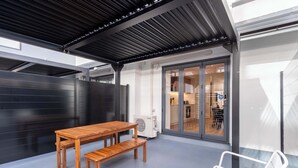 A covered outdoor seating area with a wooden table and bench, a white chair, and a glass door leading to an interior space.