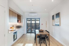 A modern kitchen with a microwave, oven, and wooden cabinets. There is a dining area with a table and chairs, and a large window with blinds. - The Ascot | 3BR + Patio | Courtenay Place (Wellington)