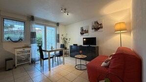 Ein Wohnzimmer mit Sofa, Fernseher, Esstisch mit Stühlen, einem kleinen Beistelltisch und einem Fenster mit Vorhängen.