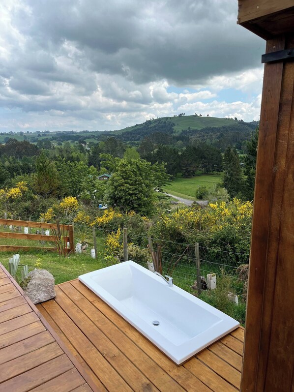 A wooden deck with a white bathtub, surrounded by greenery and a view of rolling hills.