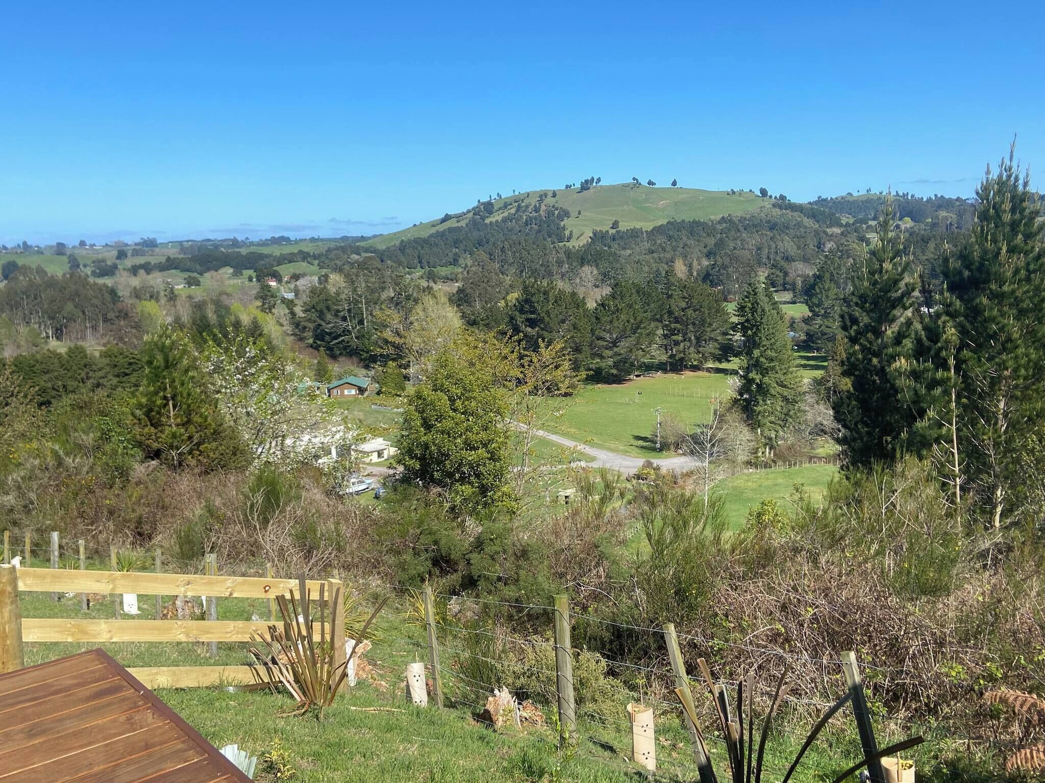 Un paysage rural avec une terrasse en bois, un espace clôturé et une vue sur une colline parsemée d’arbres.