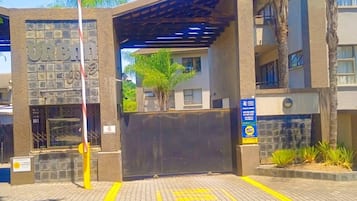 Uma entrada com portão para um edifício de fachada em pedra, galeria coberta e palmeiras.