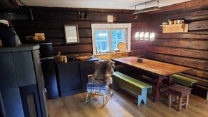 L'interno accogliente di una baita rustica con un tavolo da pranzo in legno, panche, una poltrona a dondolo e un angolo cottura con frigorifero e pensili.