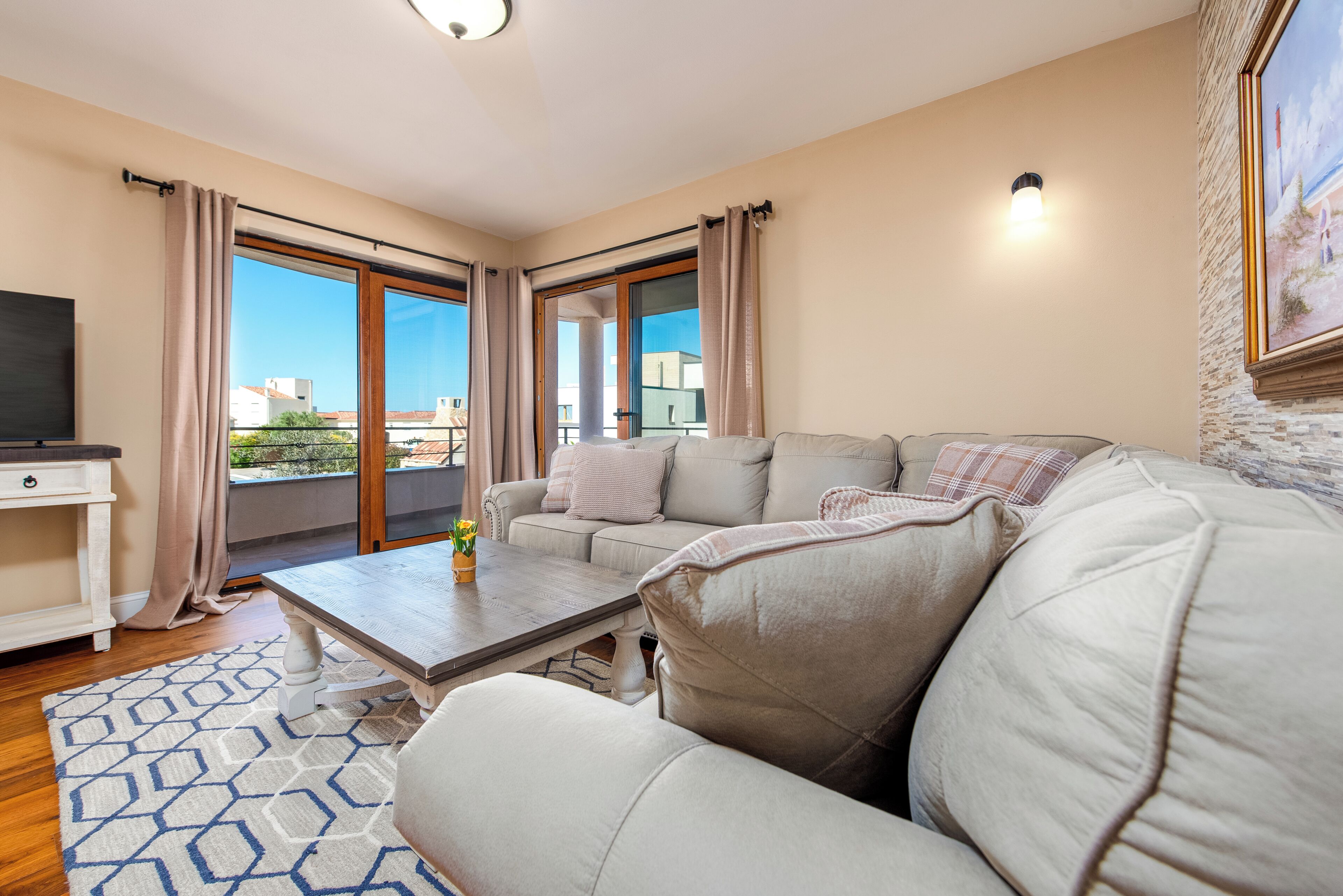 Ein Wohnzimmer mit einer Couch, einem Couchtisch und einem Fernseher. Ein Balkon mit Vorhängen und Blick auf Gebäude ist vorhanden.