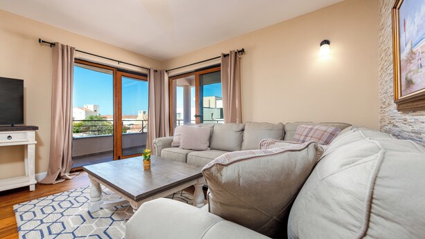 Een woonkamer met een bank, een salontafel en een televisie. Er is een balkon met gordijnen en uitzicht op gebouwen.