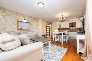 Ein Wohnzimmer mit einer Couch, einem Couchtisch und einem Bild an der Wand. Es gibt einen Essbereich mit Tisch und Stühlen, und im Hintergrund befindet sich eine Küche.
