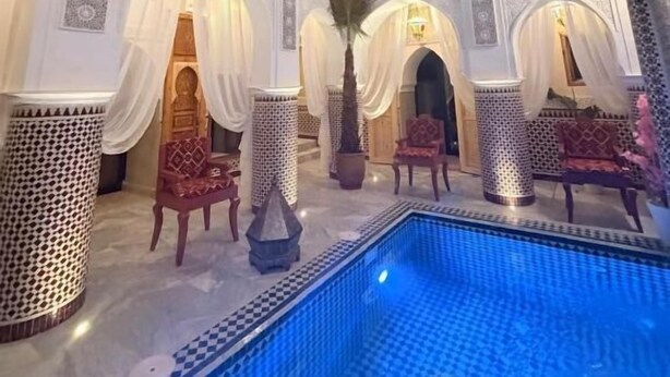 Un espace piscine intérieur luxueux, doté d’une architecture raffinée, comprenant des arches et des colonnes, et une piscine bordée d’une mosaïque bleue.