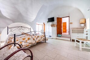 A bedroom with a curved ceiling, a bed with a floral bedspread, a wooden door, a white dresser, and a television mounted above the door. - Le Mesolecchie (Crispiano)
