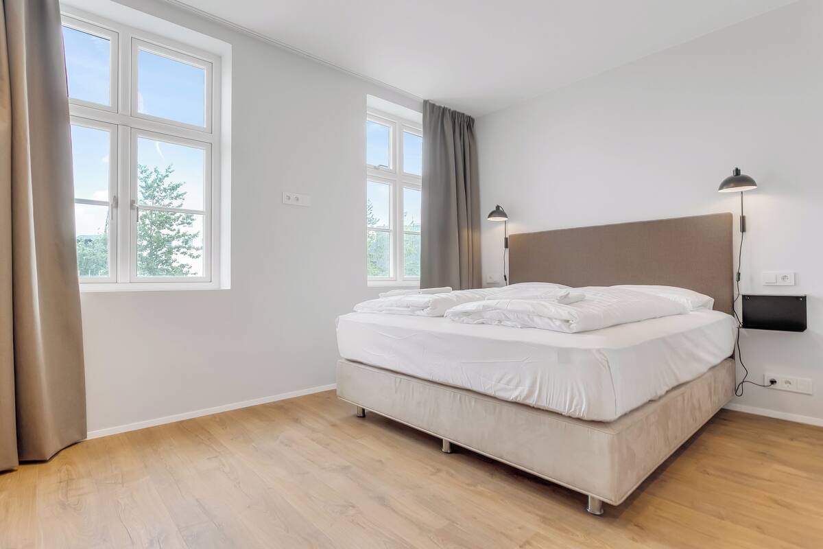 Un dormitorio con una cama, dos lámparas de noche, un ventanal y un televisor en la pared.