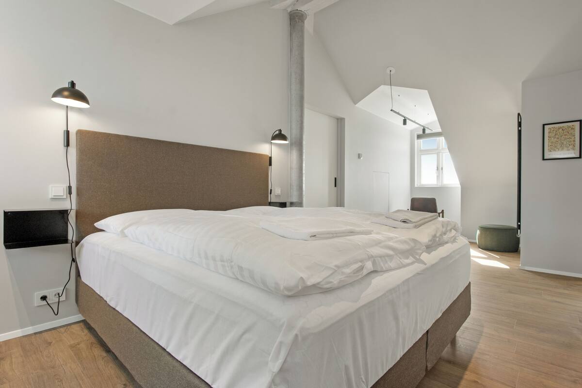 Un dormitorio moderno con una cama grande, dos lámparas de noche, piso de madera y un techo alto con tragaluces.