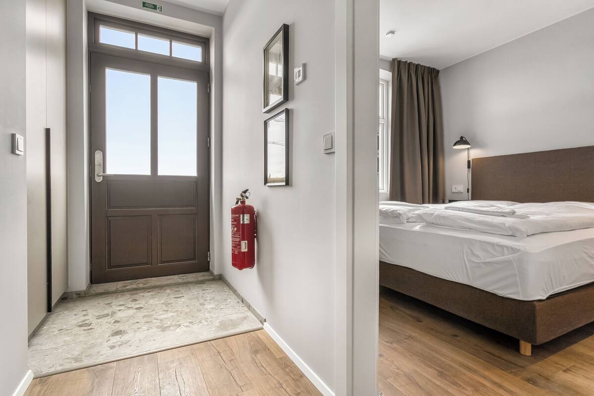 Habitación de hotel con una cama, un extintor, una puerta y una lámpara.