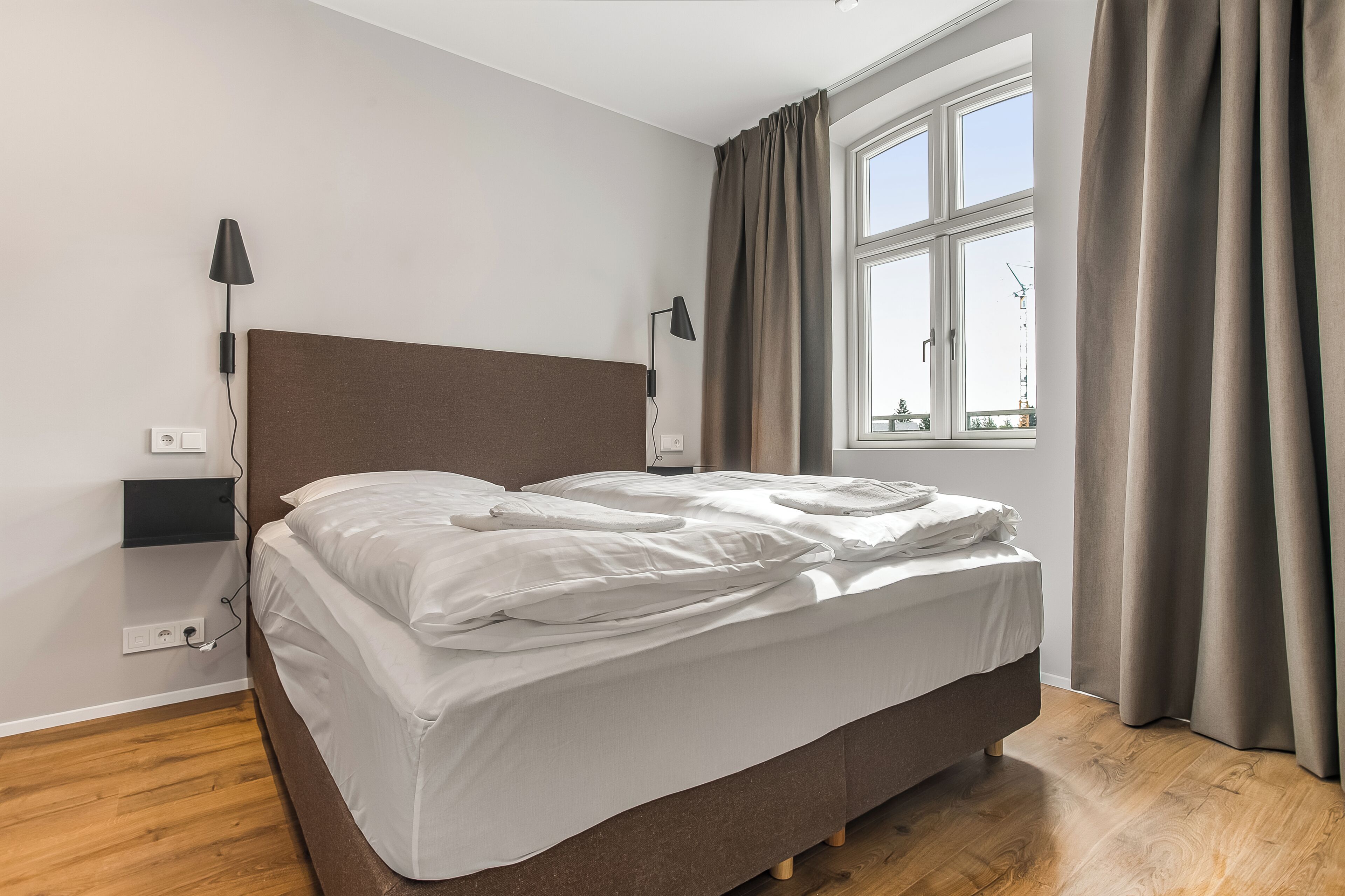A neatly made bed in a room with a window, a lamp, and a power outlet on the wall.