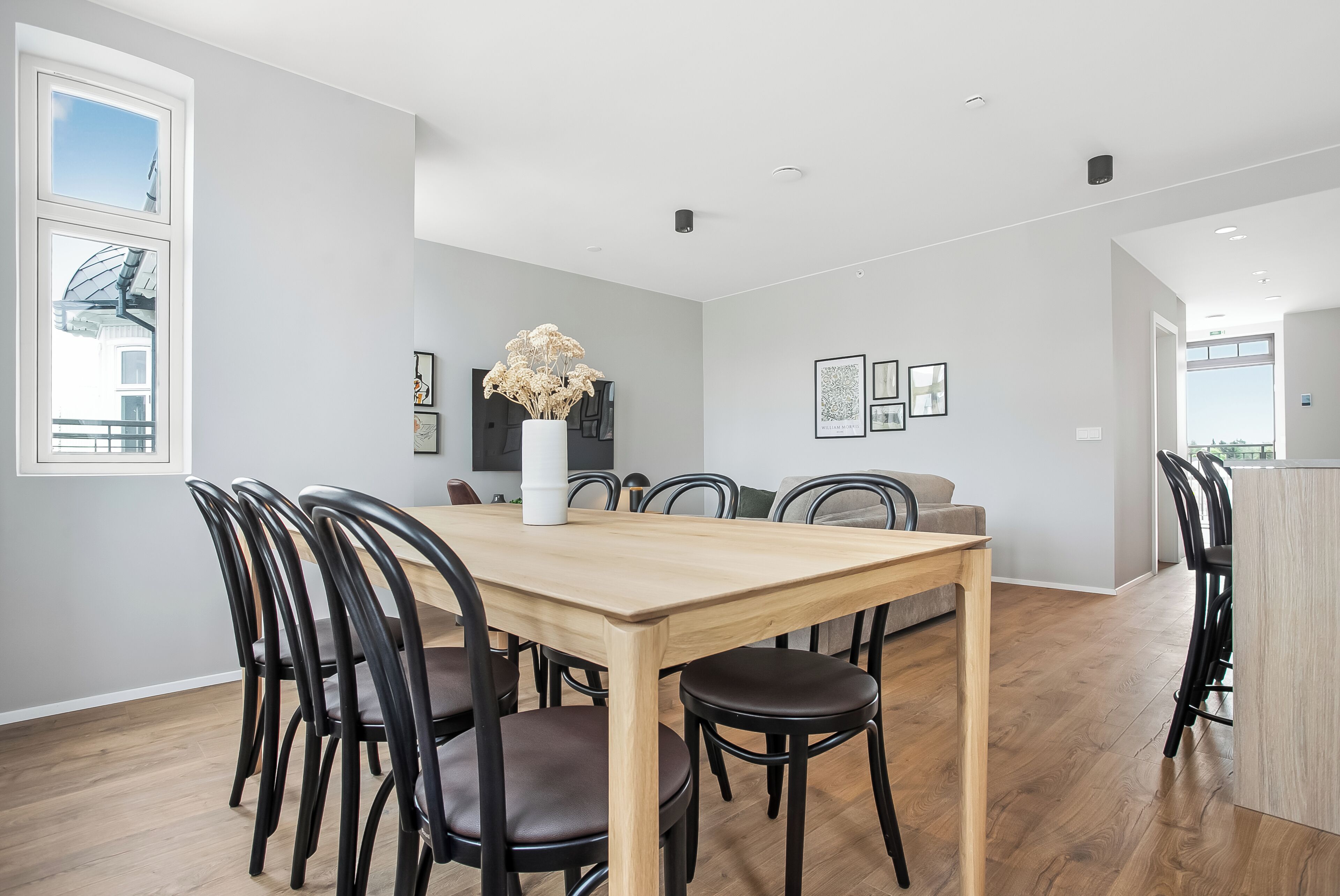 A modern dining area with a wooden table and chairs, a vase of dried flowers, and framed artwork on the wall.