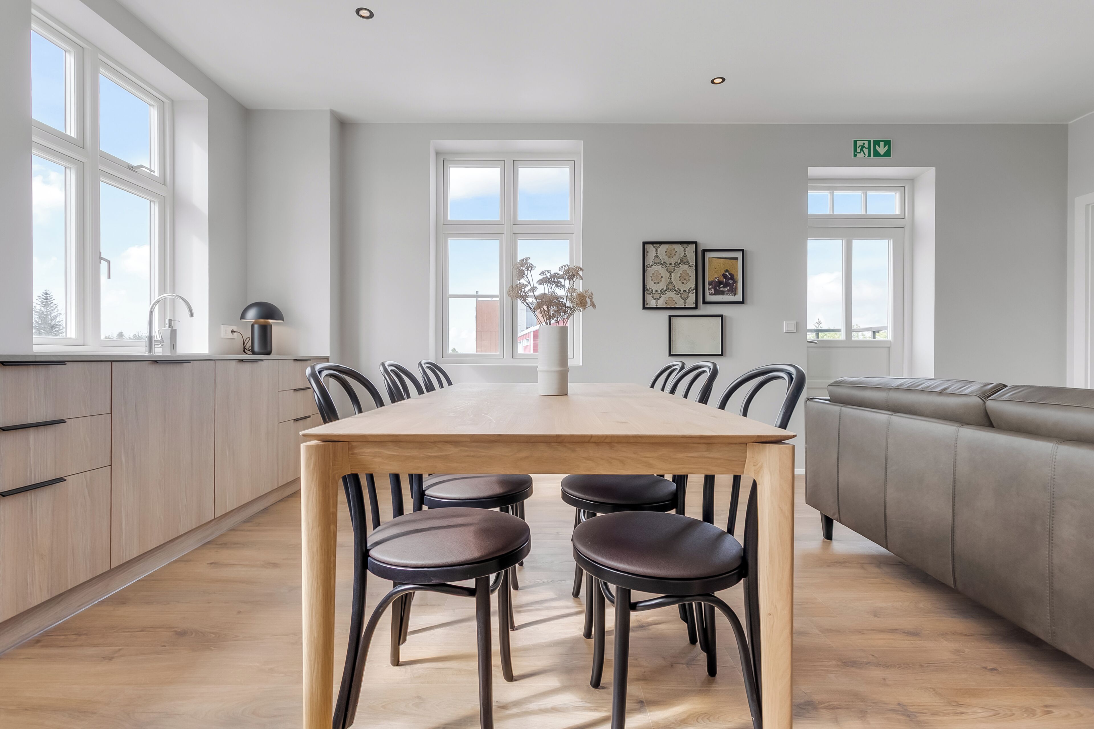 A modern kitchen with a wooden table and chairs, a grey sofa, and large windows.