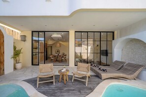 Uma área à beira da piscina com móveis de vime, uma mesa com um vaso e uma espreguiçadeira com uma pessoa deitada.