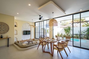 A modern interior with a dining area, a TV mounted on a stone wall, and a view of a swimming pool. - Villa Avalon Canggu (Canggu)