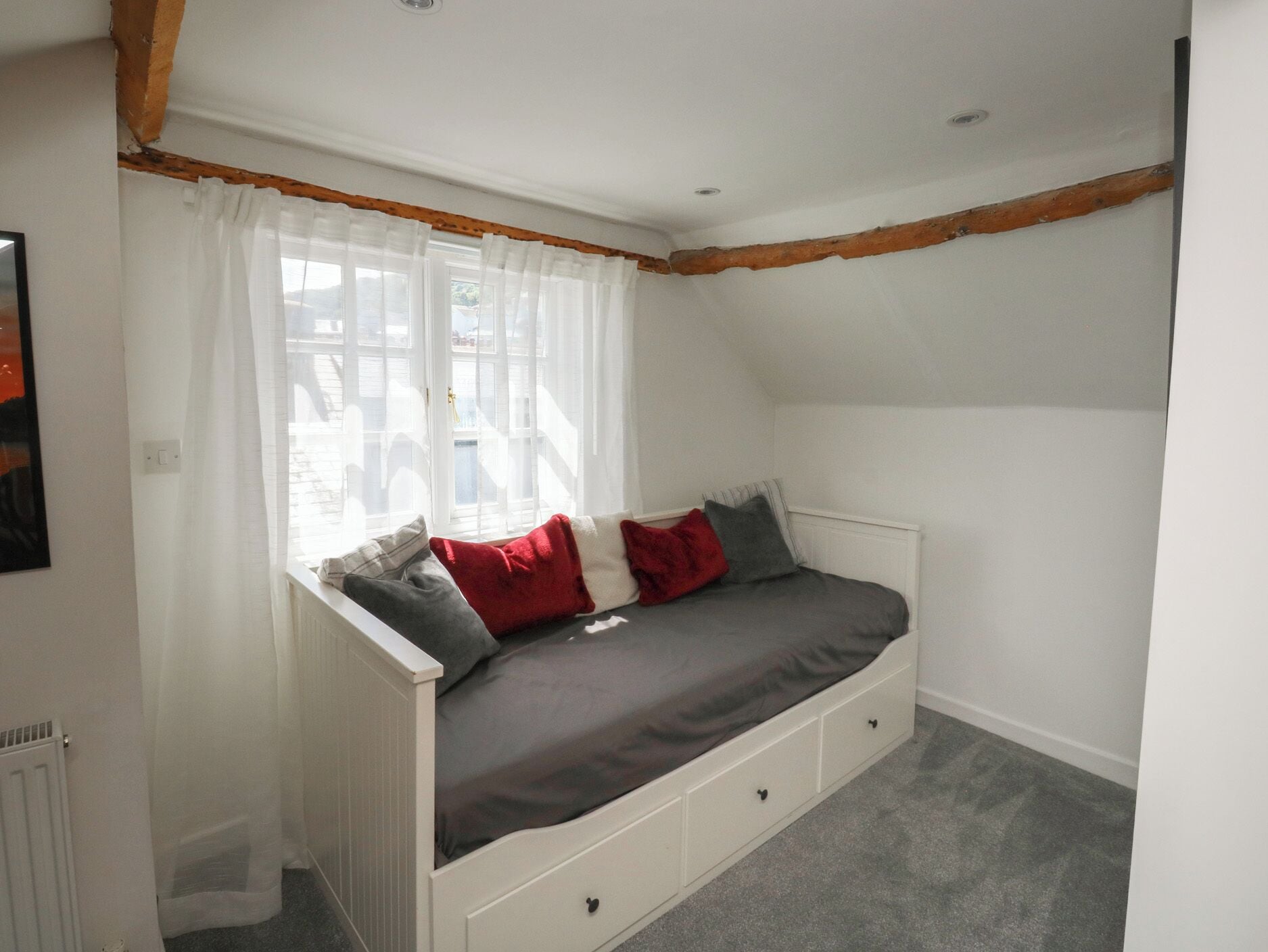 Ein Zimmer mit schrägem Dach, einem Bett mit Stauräumen, einem Fenster mit durchsichtigen Vorhängen und einer Heizung.