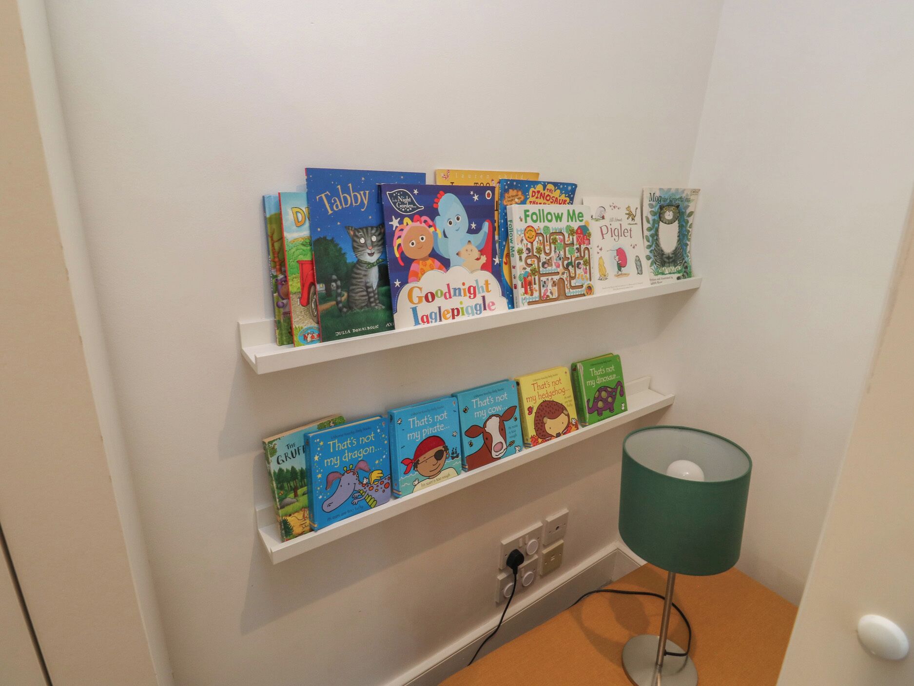 Een boekenplank met kinderboeken en een lamp op een tafel.