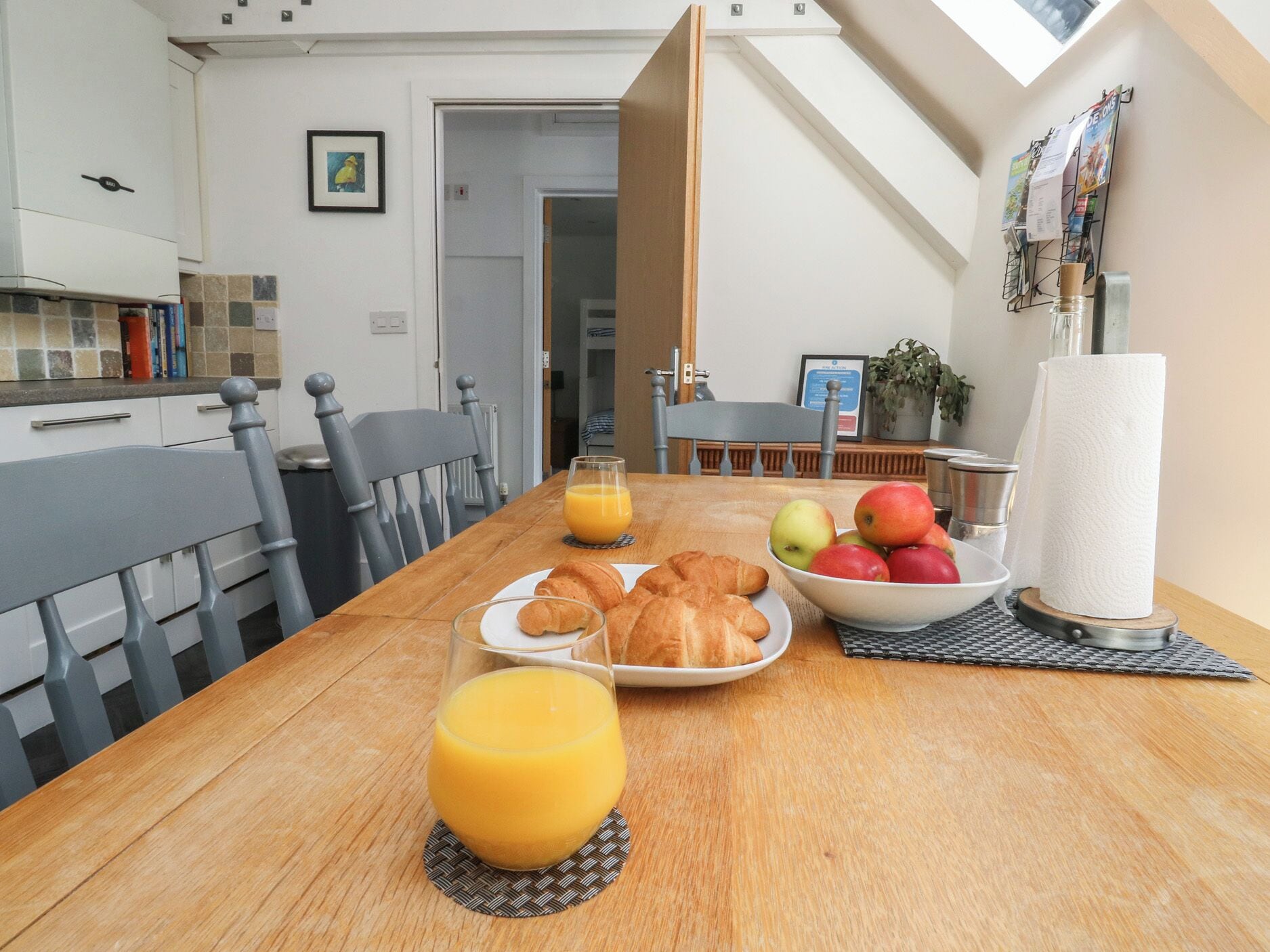 Una mesa de comedor con cruasanes, manzanas y jugo de naranja en una cocina moderna con vista al cielo.