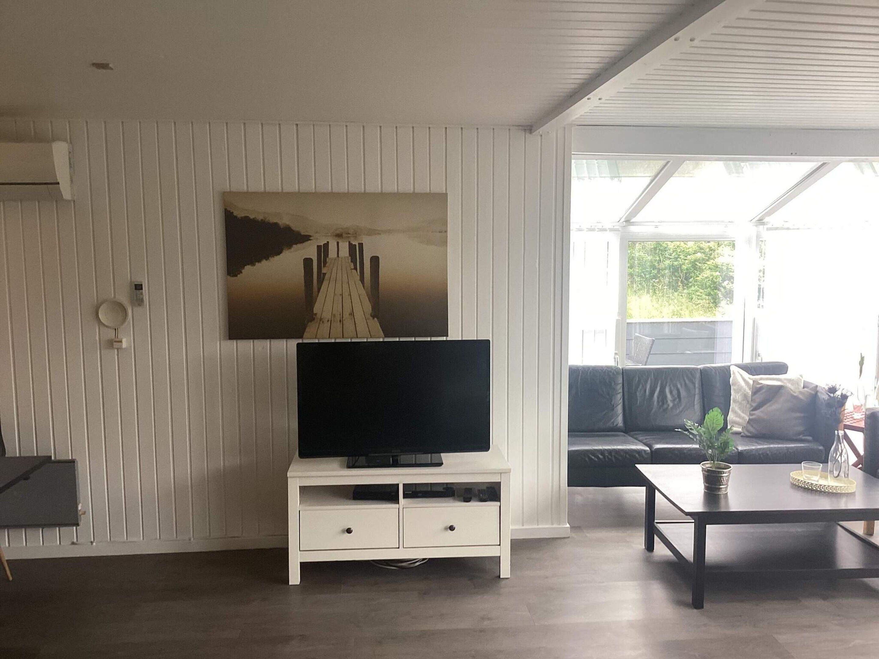 A living room with a flat-screen TV on a white stand, a sofa, a coffee table, and a window with a view of greenery.