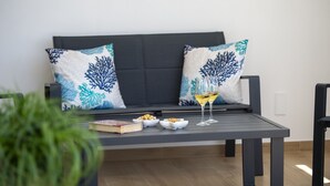 Ein dunkelgraues Sofa mit gemusterten Kissen, ein kleiner Tisch mit einem Buch, Schalen mit Snacks und ein Glas Wein.