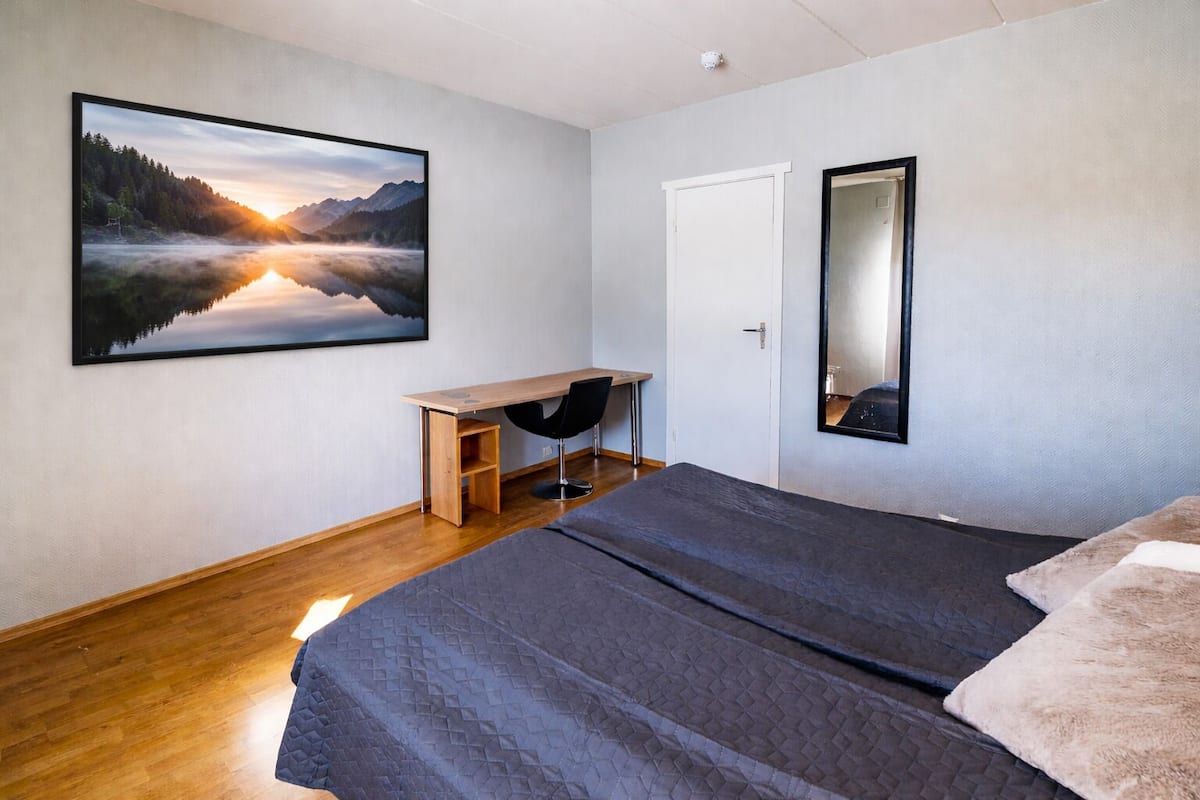 Un dormitorio con cama, escritorio, silla, un cuadro grande enmarco y un espejo.