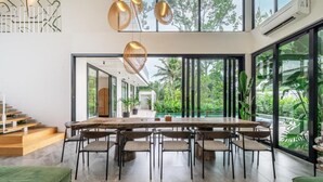 Un espace de repas moderne, doté d’une grande table et de chaises en bois, de larges fenêtres offrant une vue sur la verdure, et d’un escalier avec des marches en bois.