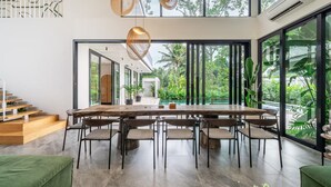 Un espace de repas moderne, doté d’une grande table et de chaises, de larges fenêtres offrant une vue sur la verdure, et d’un escalier menant à un autre niveau.