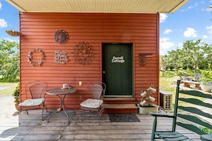 Una cabaña roja con una puerta verde que dice 'Carroll Cottage', dos sillas de metal, una mesa redonda pequeña y adornos de metal en la pared.
