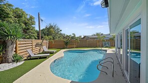 A swimming pool with a wooden fence, a lounge chair, and a glass door leading to a covered area.