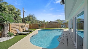 A swimming pool with a wooden fence, a lounge chair, and a glass door leading to a covered area.