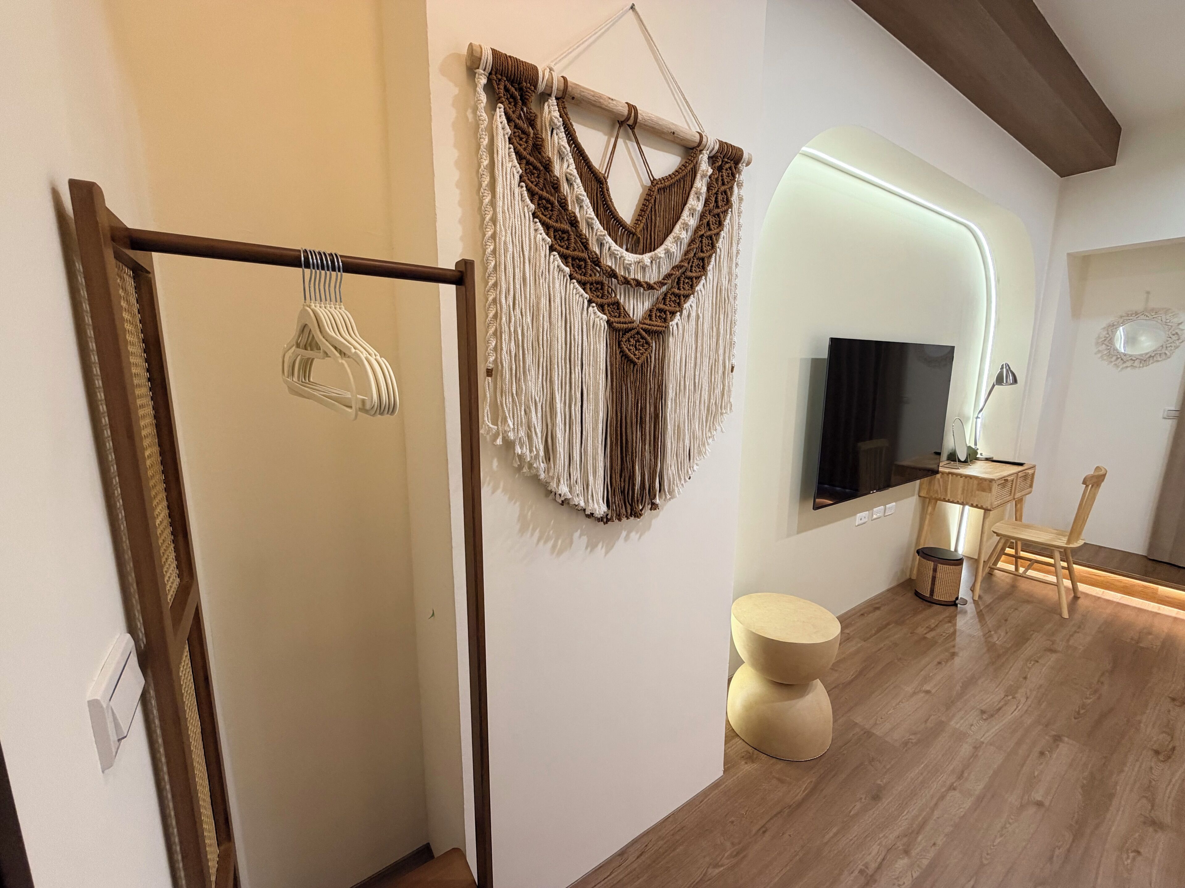 A modern room with a wooden floor, a macrame wall hanging, a flat-screen TV, a wooden chair, and a round stool.