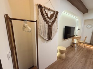 Quarto moderno com piso de madeira, um painel de macramé, TV de tela plana, uma cadeira de madeira e um banquinho redondo.