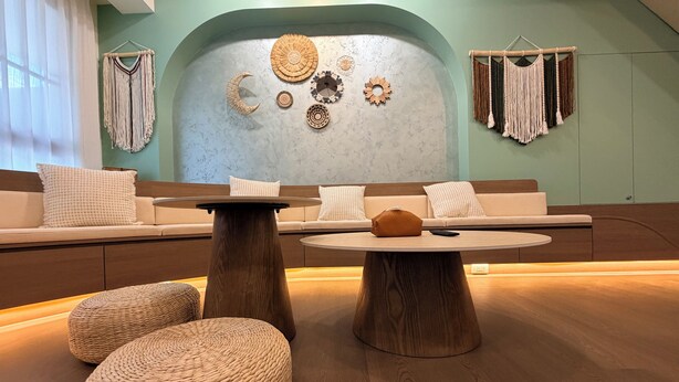 Un intérieur moderne avec une table ronde en bois, des poufs en fibres tressées, un canapé avec des coussins et des éléments de décoration murale, notamment des suspensions en macramé.