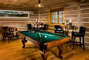 A wooden cabin interior with a pool table, chairs, a table with drinks, and a window.