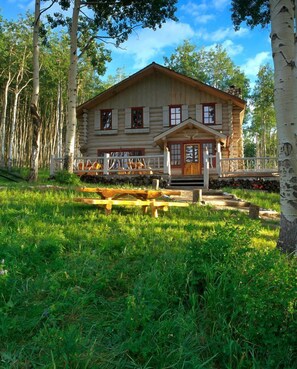 A log cabin with a front porch, surrounded by trees and a grassy area.