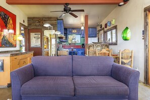 A room with a purple sofa, wooden cabinets, a ceiling fan, and a dining area with wooden chairs and a table.