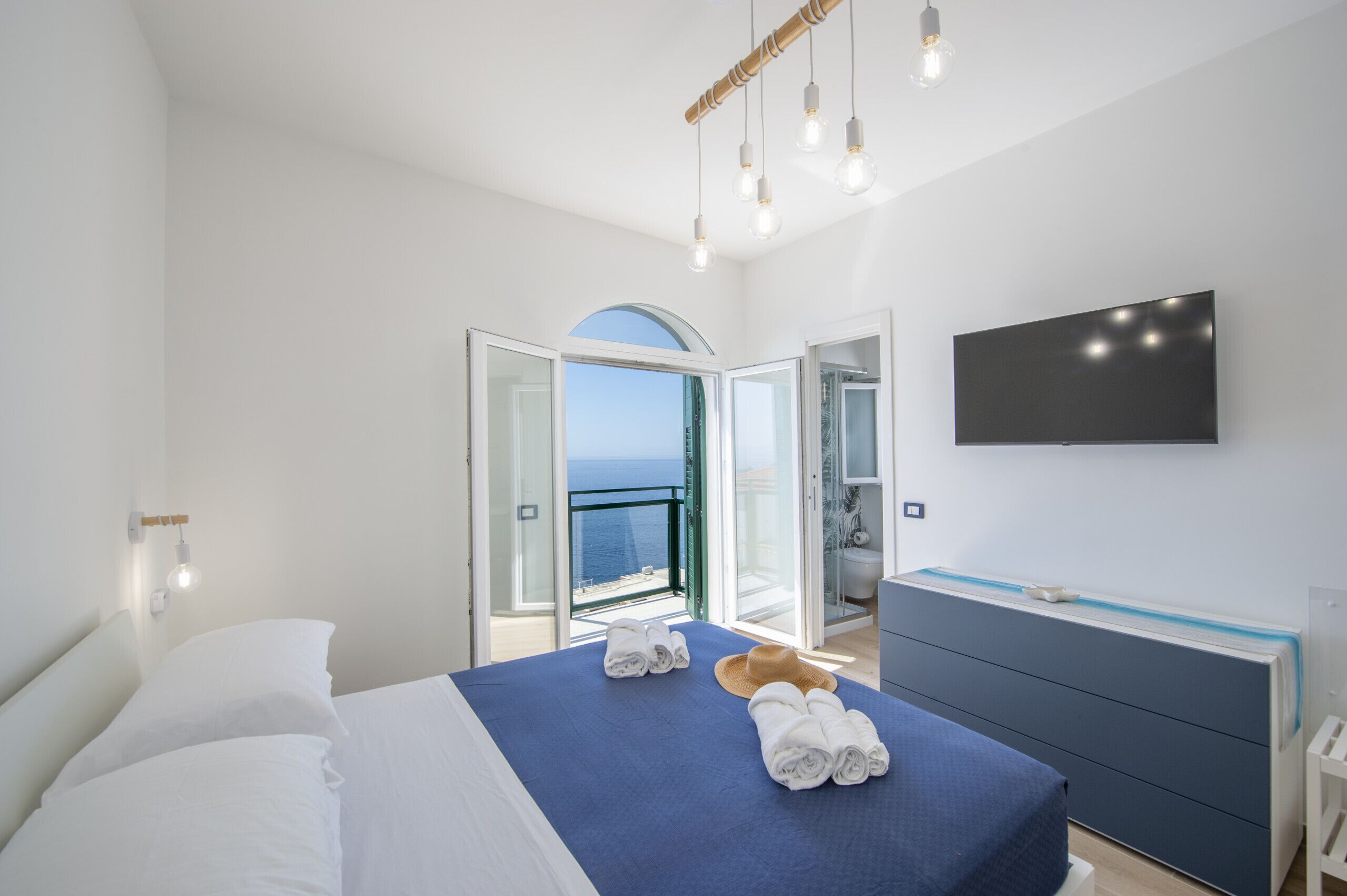 A bedroom with a bed, a TV, a dresser, and a view of the sea.