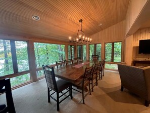 Un coin repas avec une table et des chaises en bois, de grandes fenêtres donnant sur la forêt et un lustre.