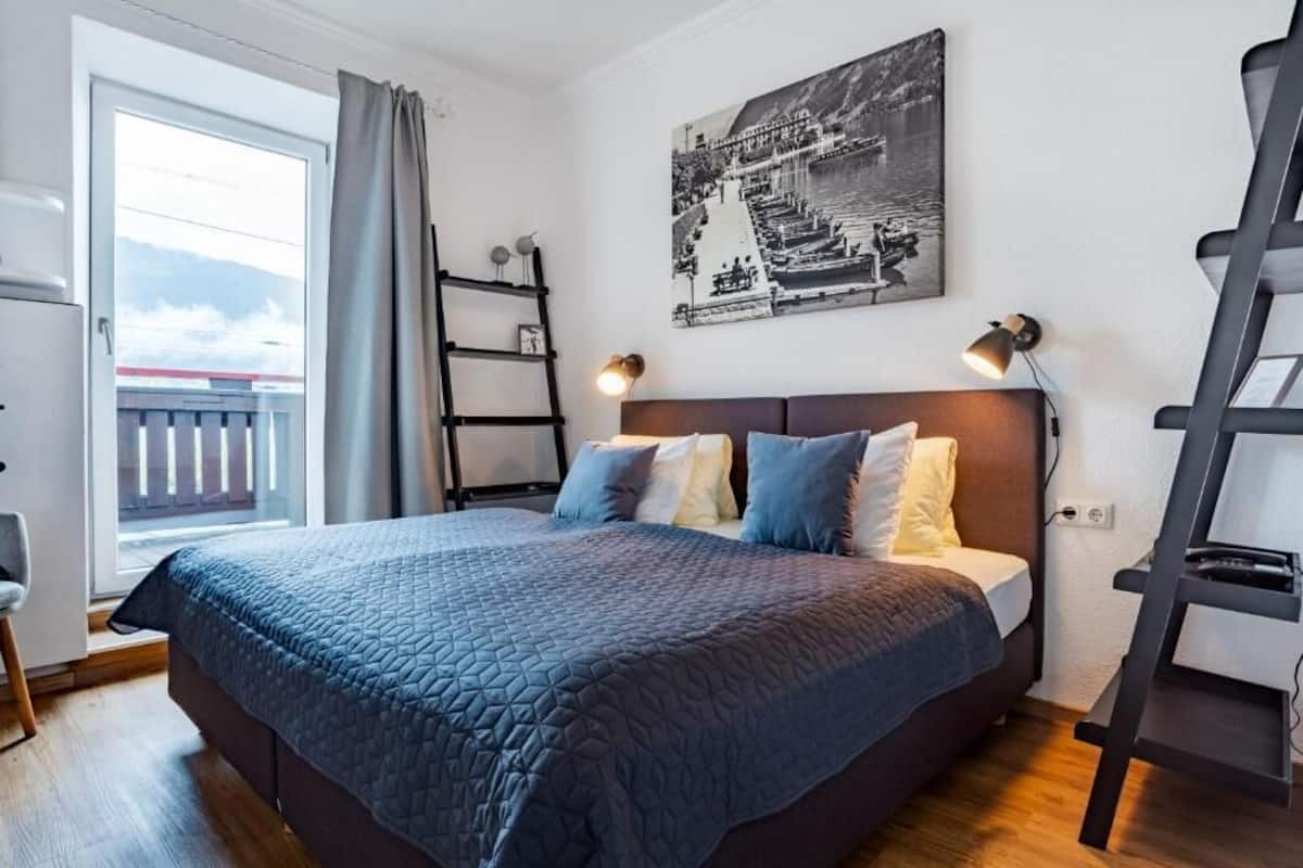 Ein Schlafzimmer mit Bett, Fenster mit Vorhängen, einer Leiter und einem Wandbild.
