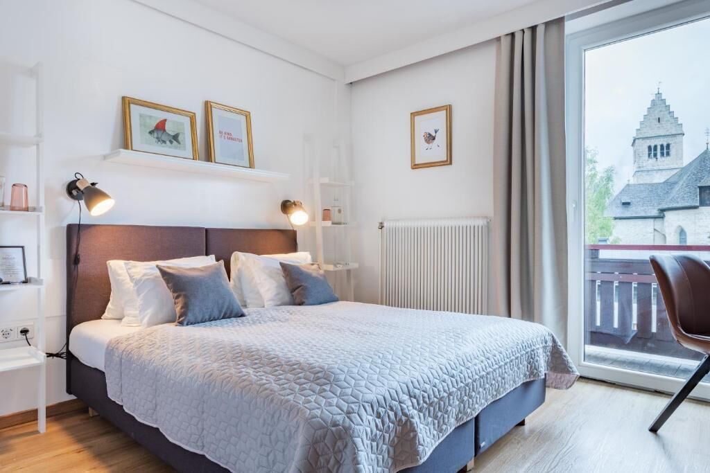 A bedroom with a bed, bedside lamps, a window with a view of a building, and a chair.