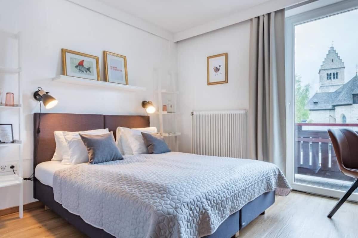 Ein Schlafzimmer mit Bett, Nachttischlampen, einem Fenster mit Blick auf ein Gebäude und einem Sessel.