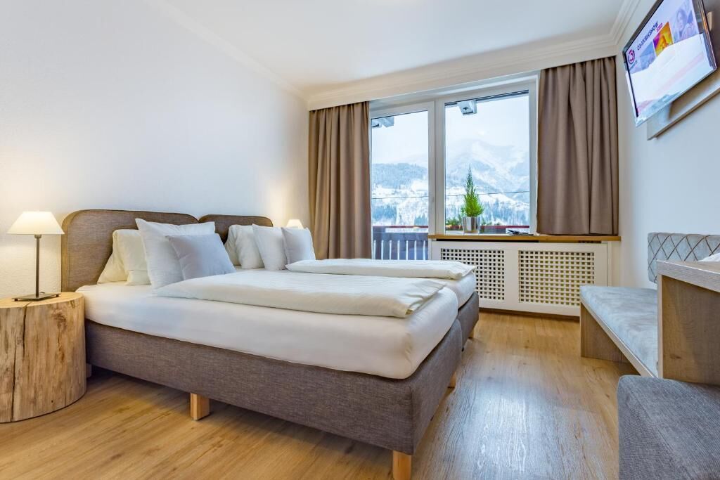 Quarto de hotel com cama, luminárias de cabeceira, vista para montanhas, janela com cortinas e uma pequena mesa com uma planta.
