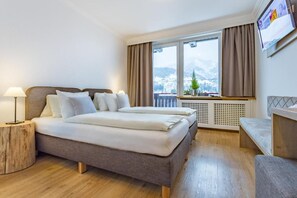A hotel room with a bed, bedside lamps, a view of mountains, a window with curtains, and a small table with a plant. - Lakeside Hotel SEEHOF I Lounge & Living (Zell am See)