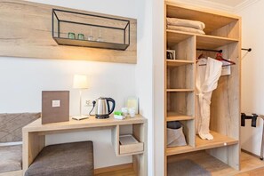 A compact hotel room with a wooden desk, a lamp, a kettle, and a built-in wardrobe with hanging clothes. - Lakeside Hotel SEEHOF I Lounge & Living (Zell am See)