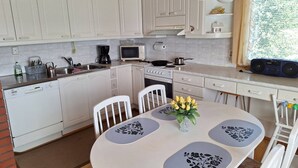 A kitchen with white cabinets, a dining table with floral placemats, and a window with curtains. - Sun's Tupa - Traveller's Marmalade (Siikalatva)