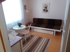 Pieni, viihtyisä huone, jossa on yksi sänky, valkoinen tuoli, pieni pöytä, jossa on maljakko täynnä kukkia, ja raidallinen matto.