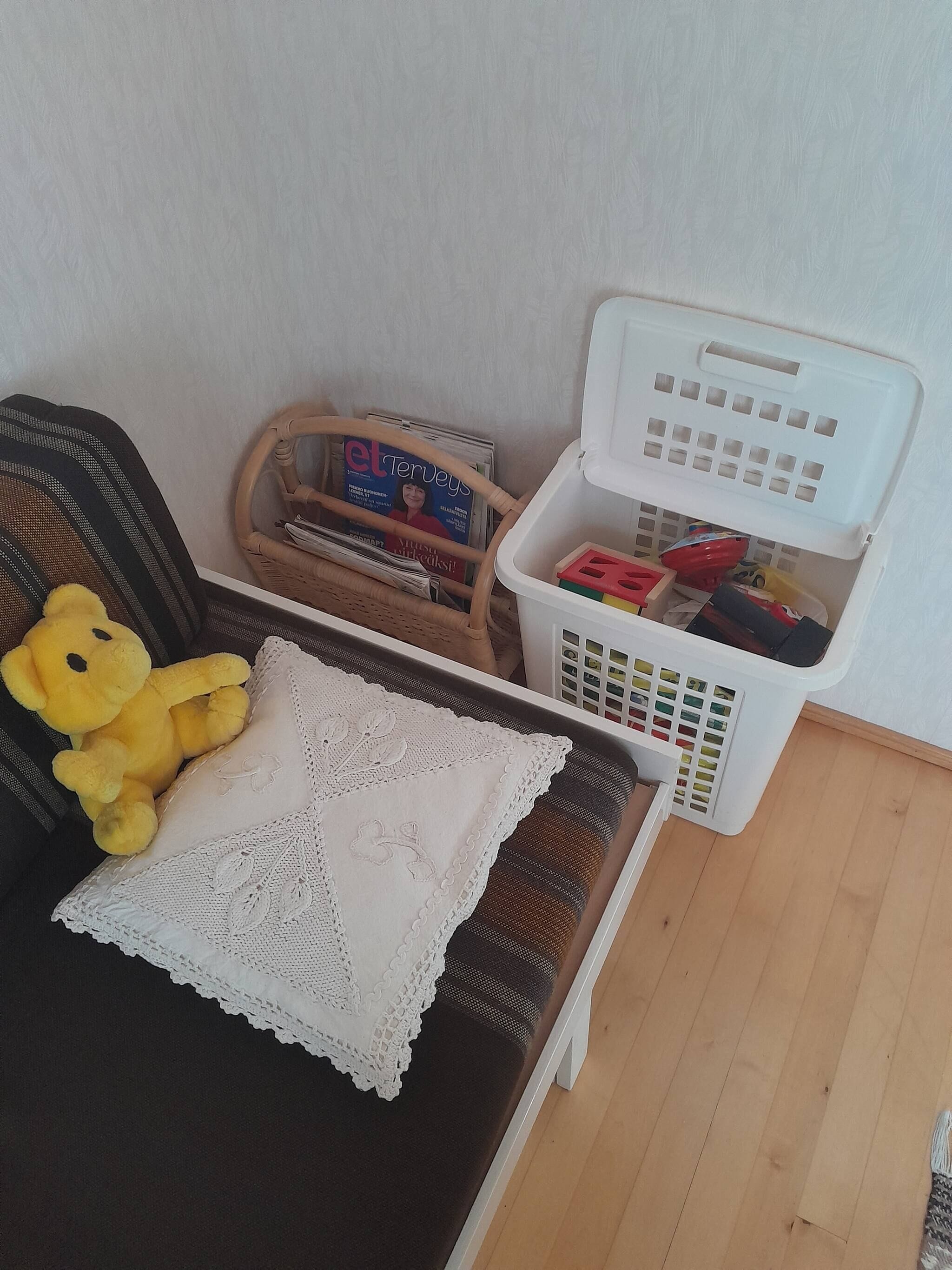 Et lille rum med en fletstol, en gul teddybjørn, et magasinholder og en hvid vasketøjskurv.